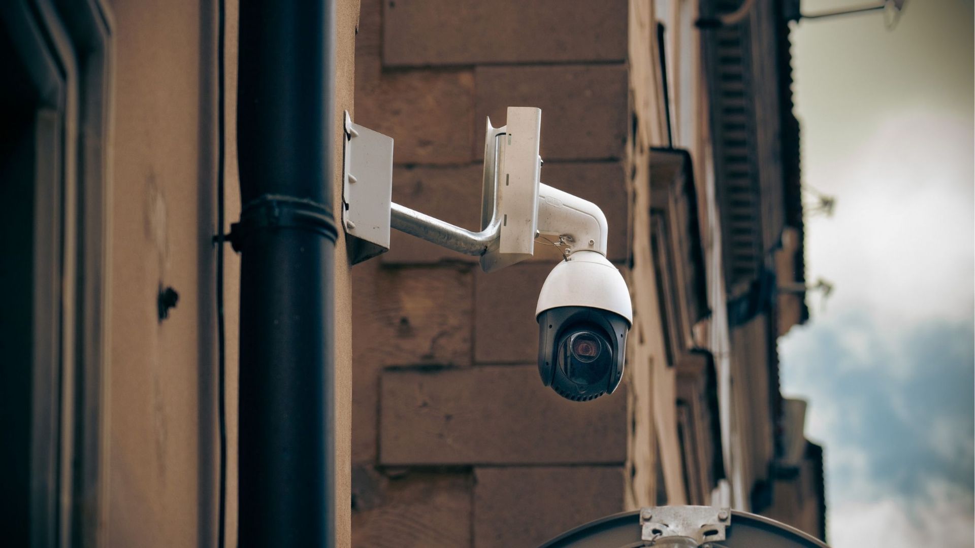 Security camera mounted on a brick building, white and black dome, bracket, cloudy sky.