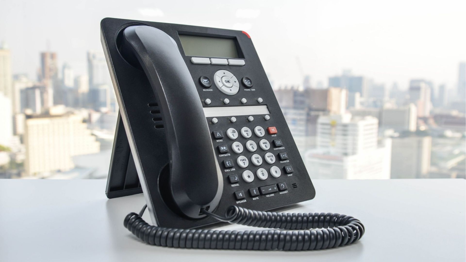 Black office telephone with a coiled cord, on a white desk with city skyline in the background.