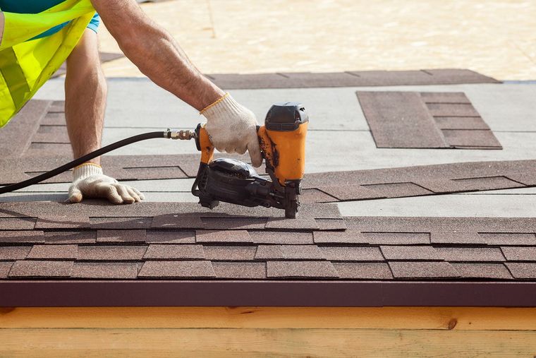 Worker using a nail gun to install brown shingles on a rooftop Worker using a nail gun to install brown shingles on a rooftop