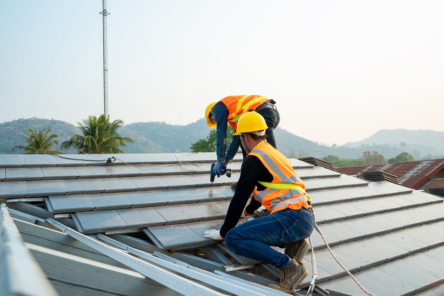 Two workers in yellow hard hats installing metal roofing on a sloped rooftop outdoors. Two workers in yellow hard hats installing metal roofing on a sloped rooftop outdoors.