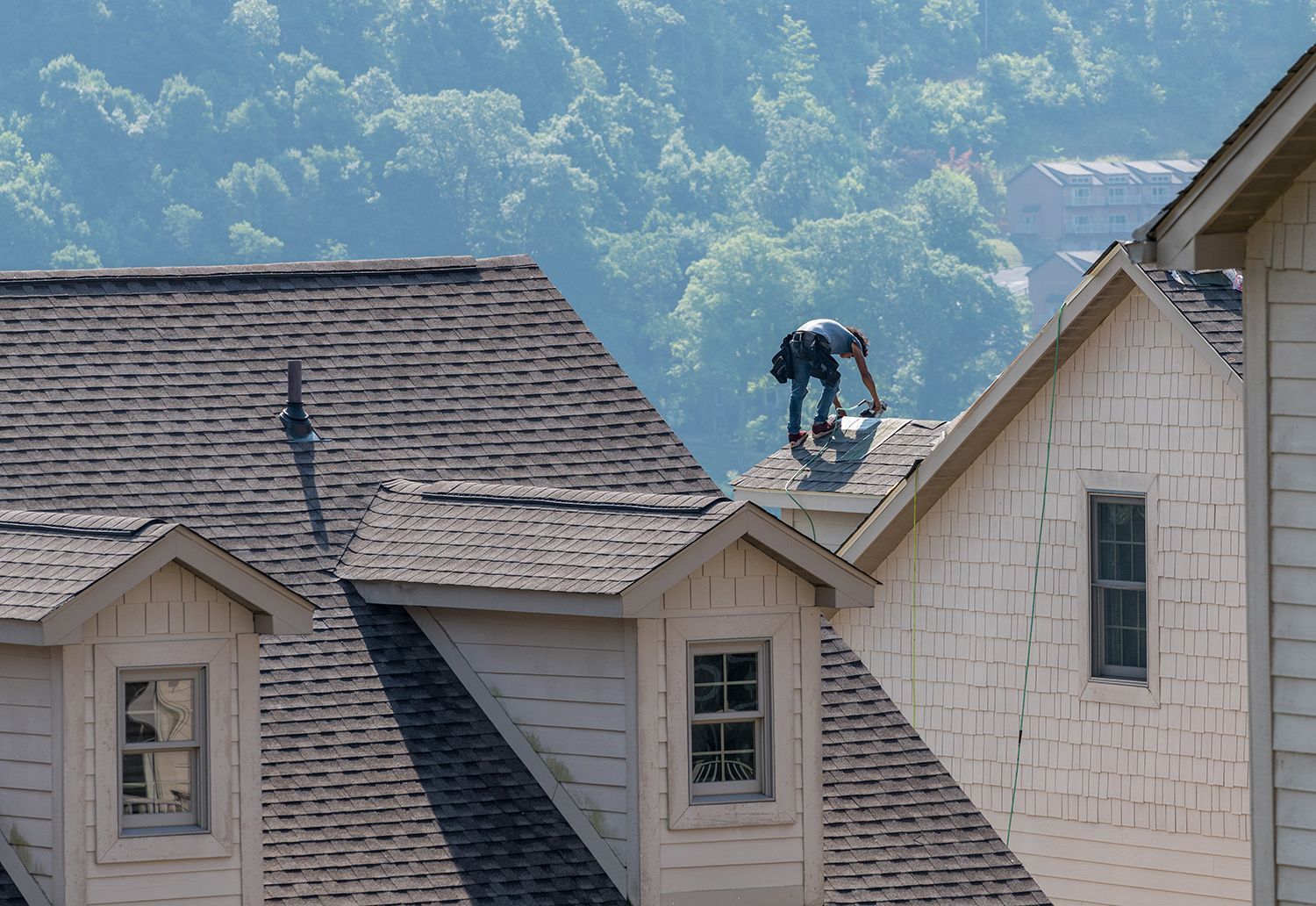 Worker on a rooftop repairing shingles on a beige house, with forested hills in the background Worker on a rooftop repairing shingles on a beige house, with forested hills in the background
