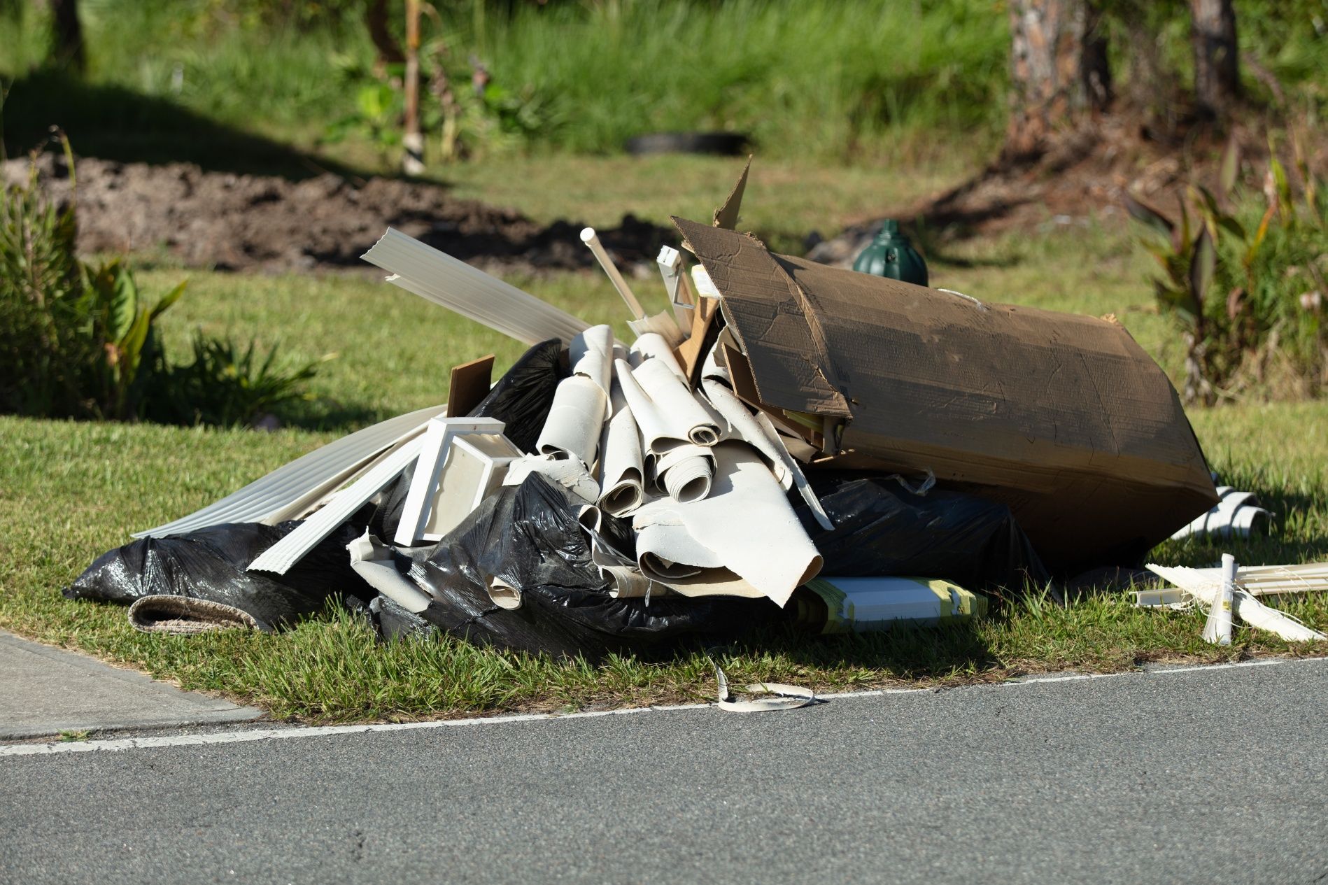 A pile of construction debris and bagged waste sits on a grassy curb next to a road.
