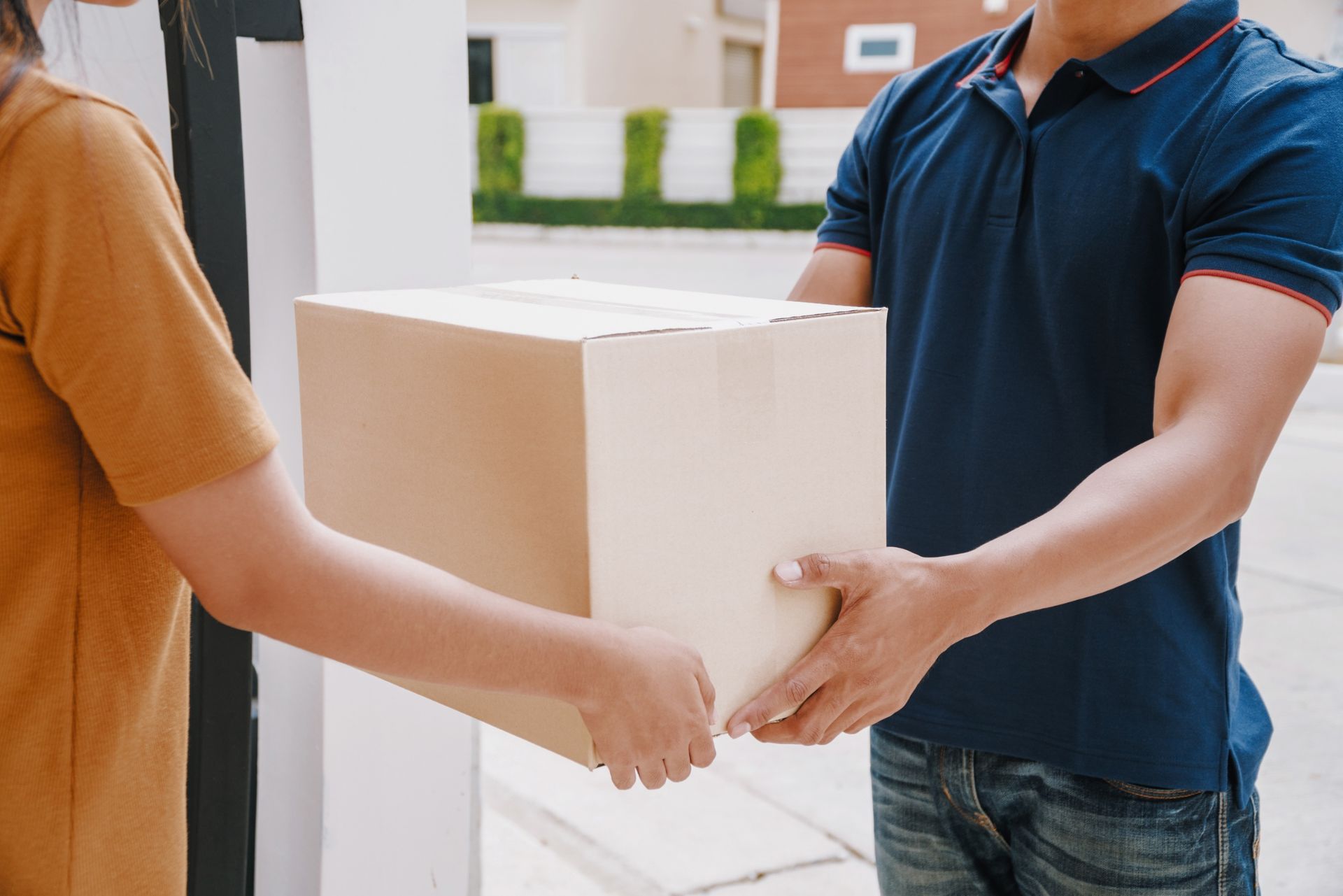 A delivery worker in a blue polo shirt hands a cardboard box to a person in an orange shirt.