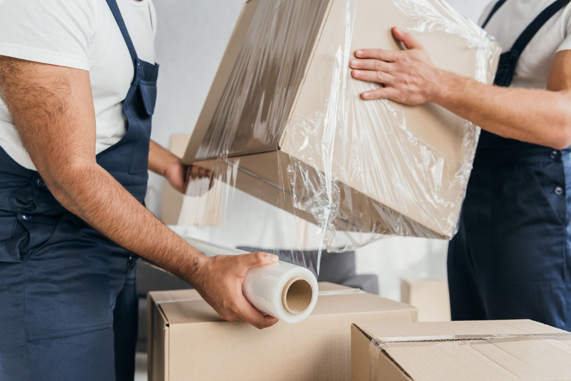 Two movers in dark blue uniforms wrap a cardboard box in clear plastic film.