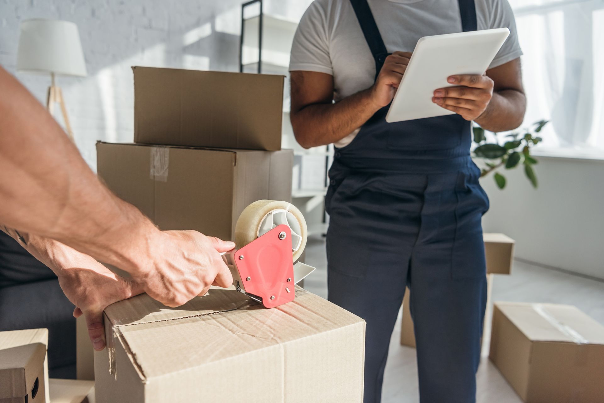 A person seals a cardboard box with a tape dispenser while another person checks a tablet in a room filled with boxes.