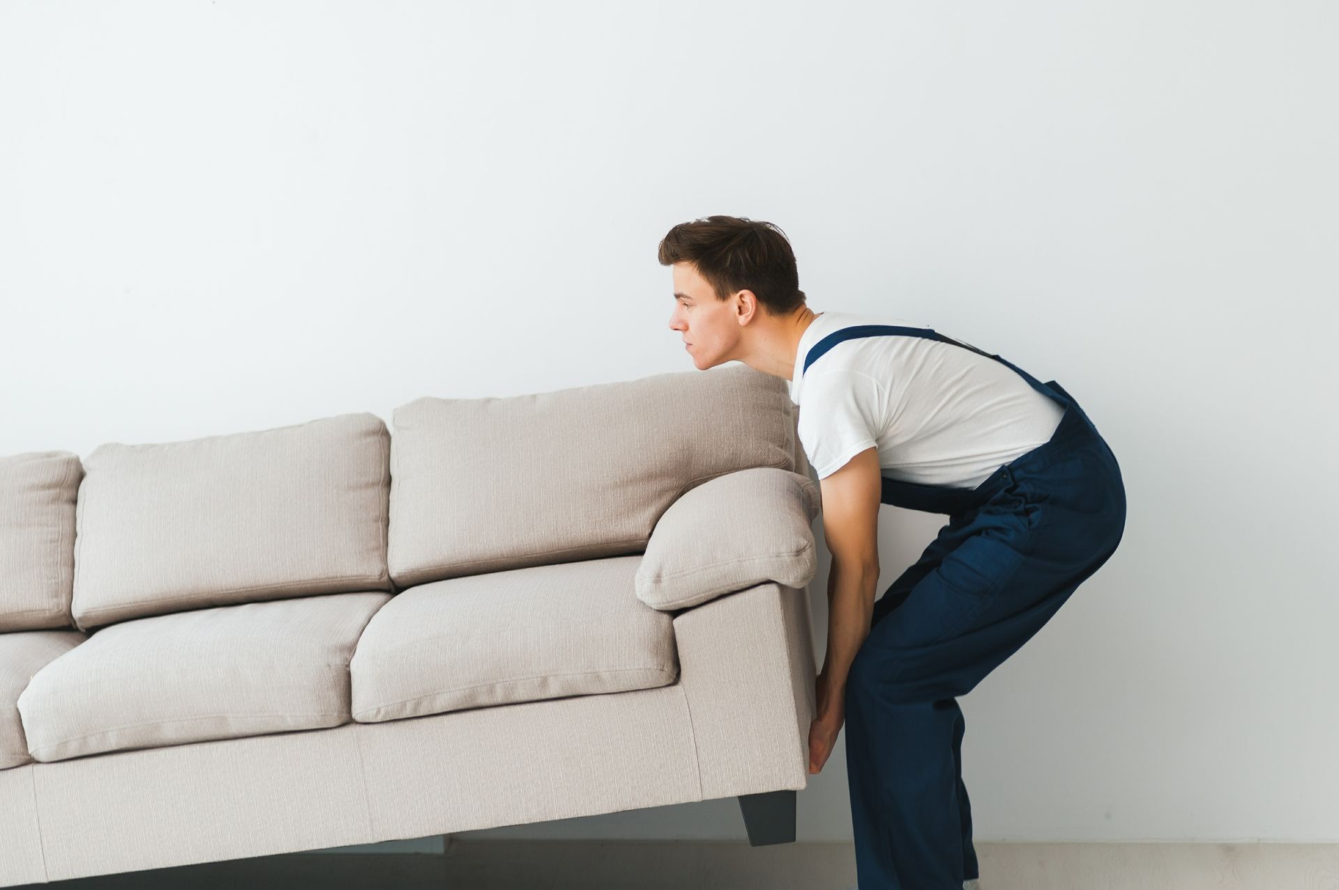 A person in blue overalls lifts one side of a beige sofa against a plain white wall.