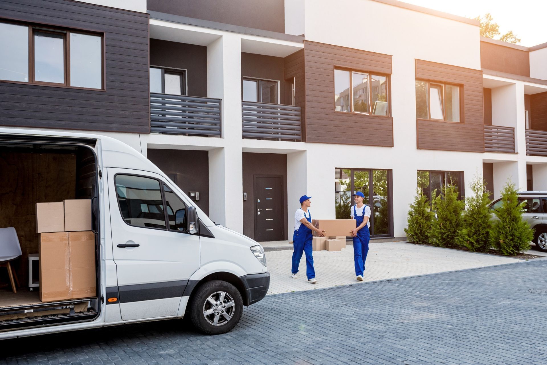 Two workers in blue uniforms carry a cardboard box toward a white moving van parked in front of modern townhouses.