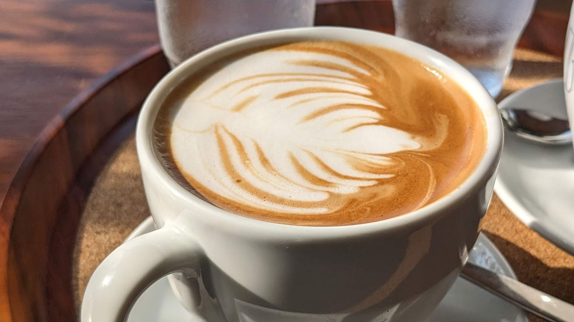 A cup of cappuccino with a leaf design on the foam is on a wooden tray.