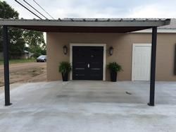 Building entrance with black doors, covered by a gray metal awning supported by black posts.