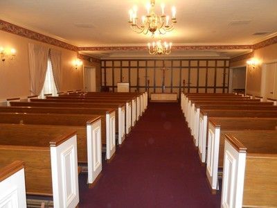 Interior of a funeral home chapel with rows of wooden pews, a red carpet, and a chandelier.