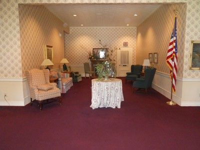 Funeral home interior with chairs, table, television, American flag. Red carpet, floral wallpaper.
