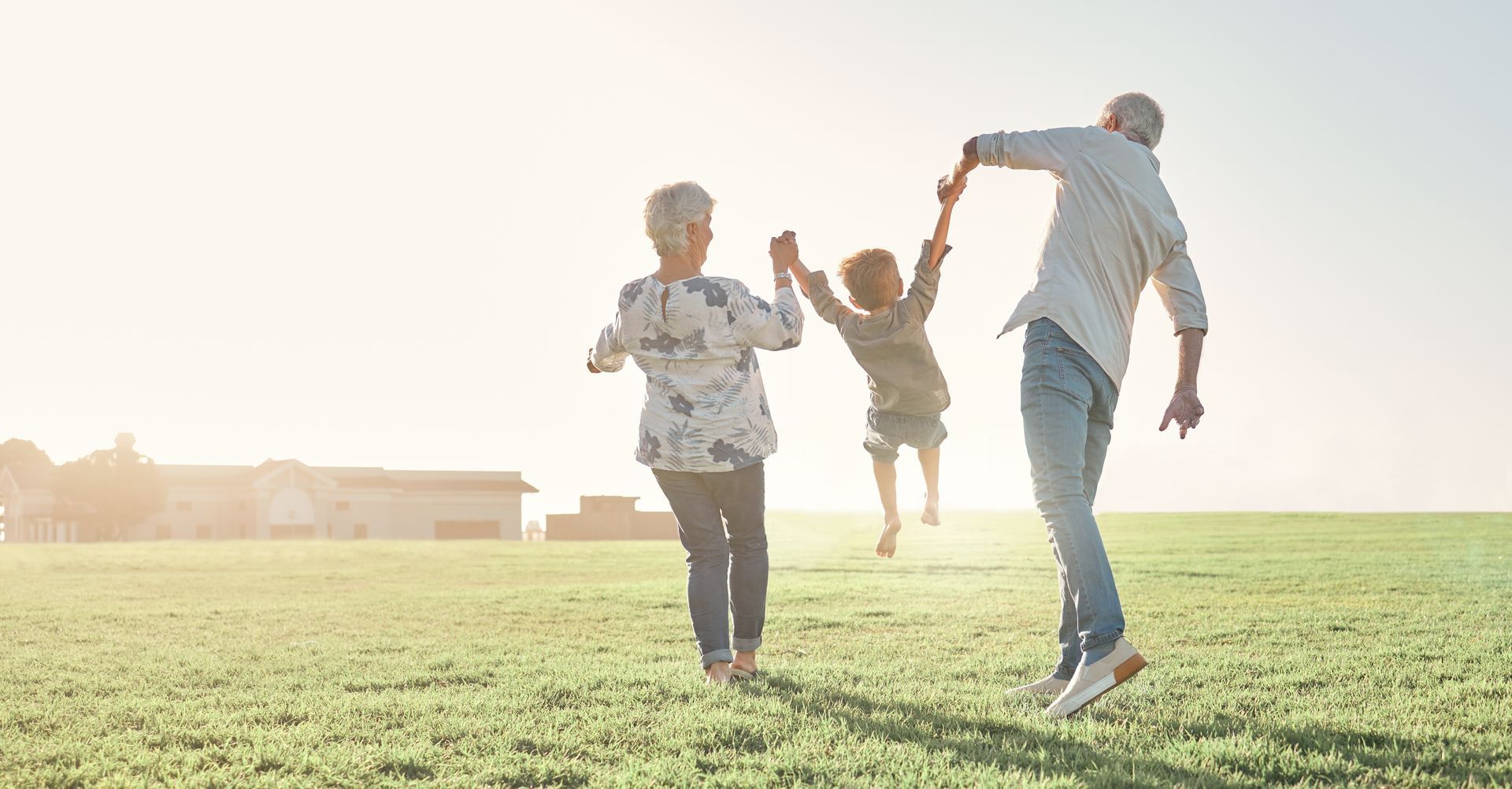 Grandparents swinging a child in a sunny field, laughing, outdoors.