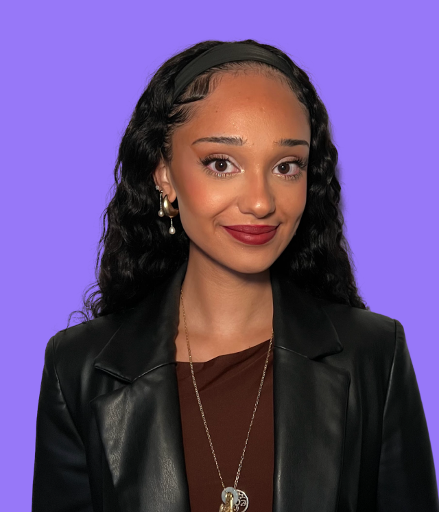 A woman wearing glasses and a blue shirt is standing in front of a purple background.