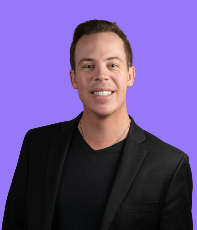 A man in a black suit and black shirt is smiling in front of a purple background.
