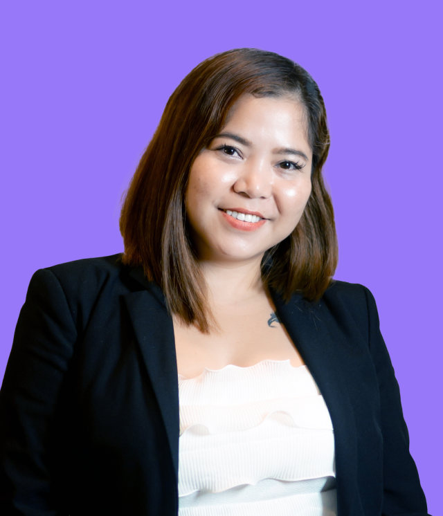 A woman in a black jacket and white top is smiling against a purple background.