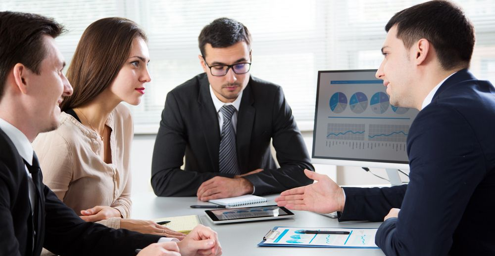 Cuatro profesionales vestidos de traje colaboran alrededor de un escritorio con gráficos y un monitor de ordenador en una oficina luminosa.