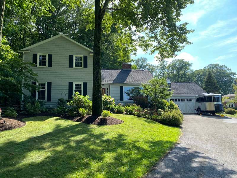 Two-story light green house with black shutters, a dark roof, and a driveway, set among trees on a sunny day.
