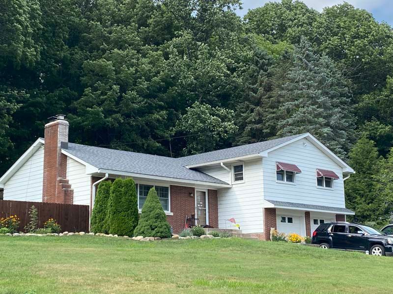 Two-story house with white siding, brick accents, a dark roof, and a green lawn. Forest in the background.