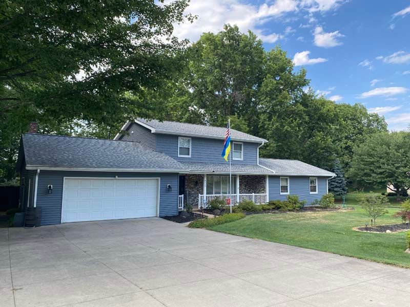 Blue house with white garage door and long driveway, surrounded by green trees and grass under a blue sky.
