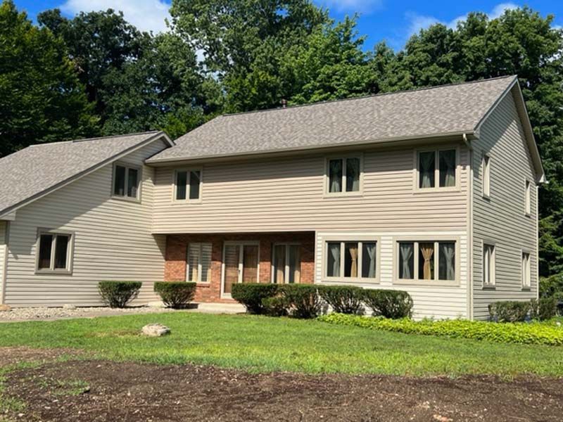 Two-story beige house with brown roof; green lawn and trees in background.