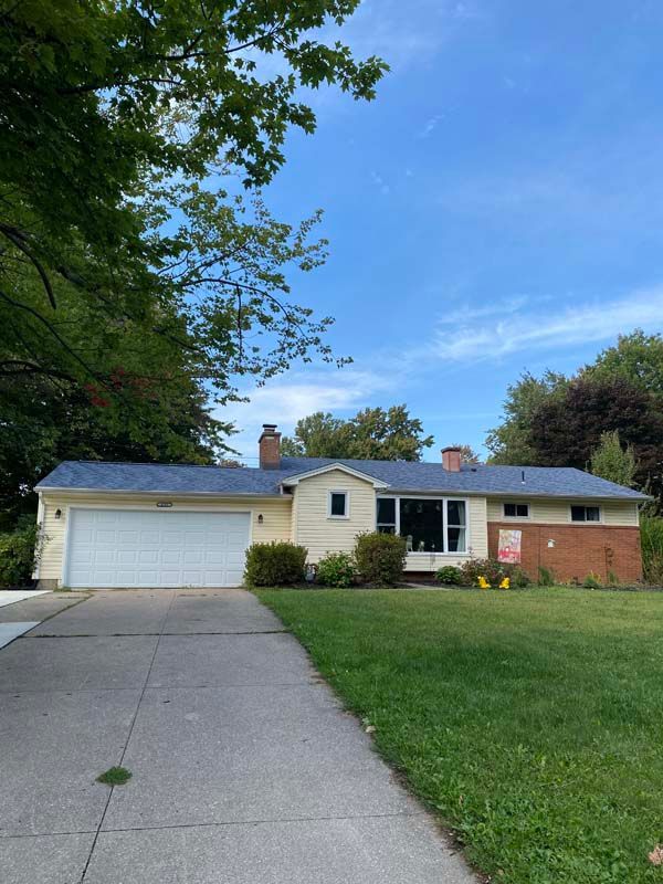 Yellow house with white garage door and brick accent, driveway, and green lawn under a cloudy sky.