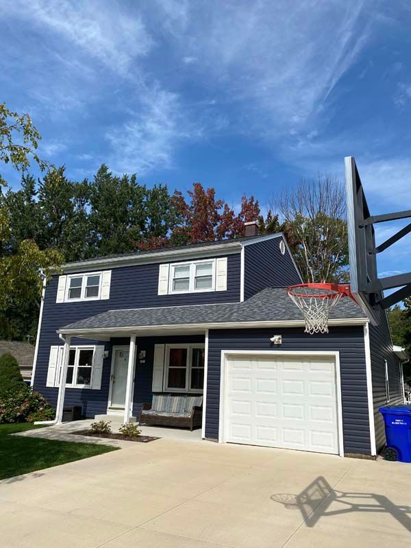 Blue-sided house with basketball hoop on a sunny day. White garage door, shutters, and concrete driveway.