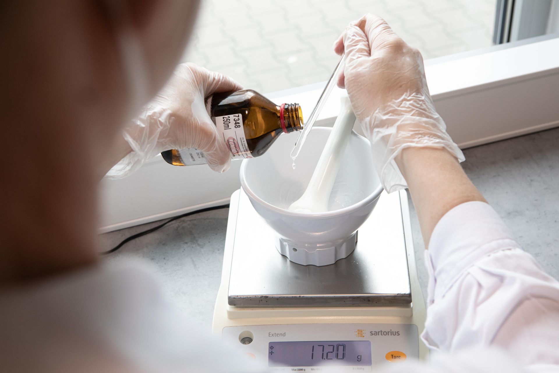 Person mit Handschuhen gießt Flüssigkeit aus einer braunen Flasche in einen Mörser auf einer Waage.