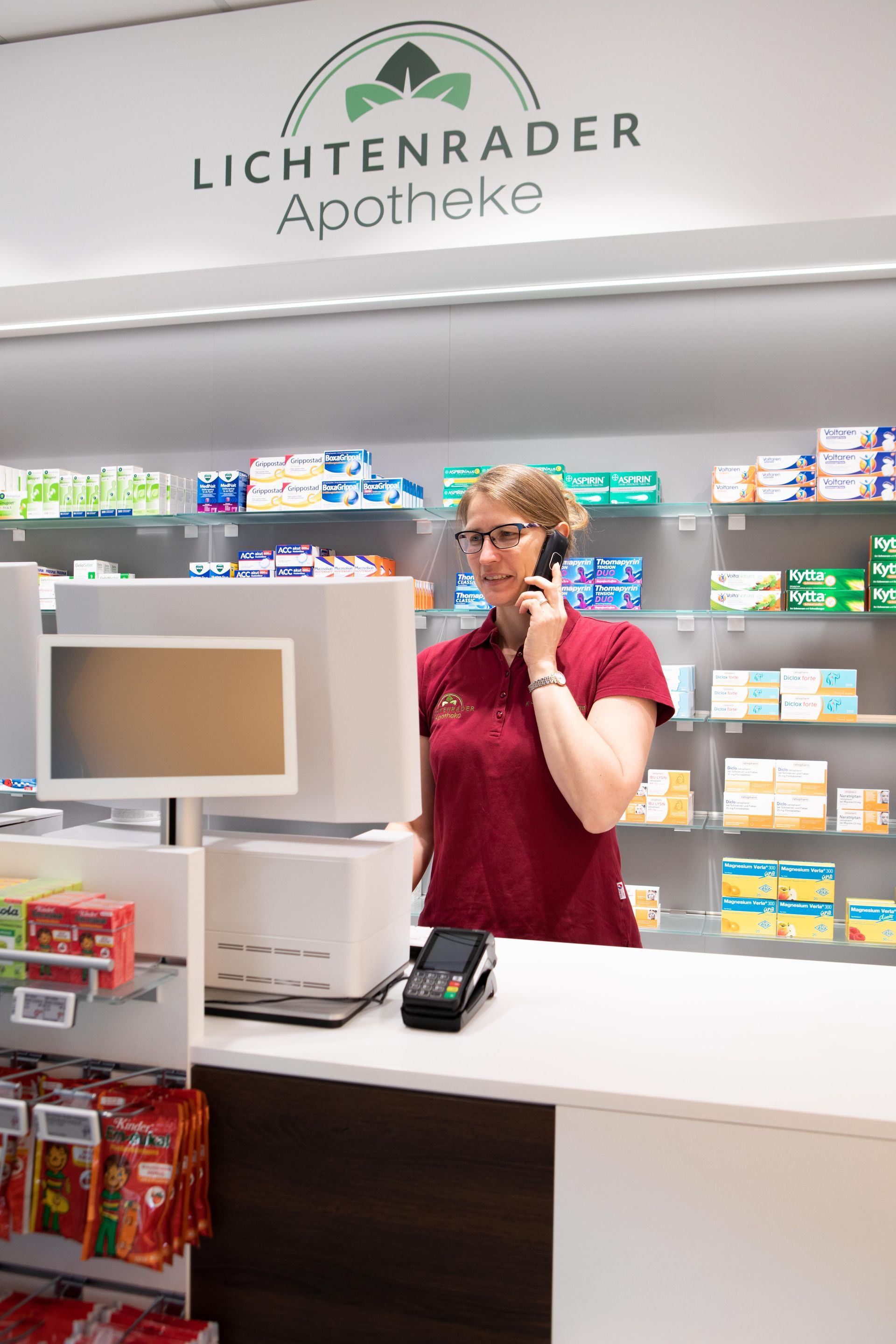 Ein Apotheker nimmt an einem Apothekenschalter einen Anruf entgegen. Im Hintergrund Regale mit Medikamenten.