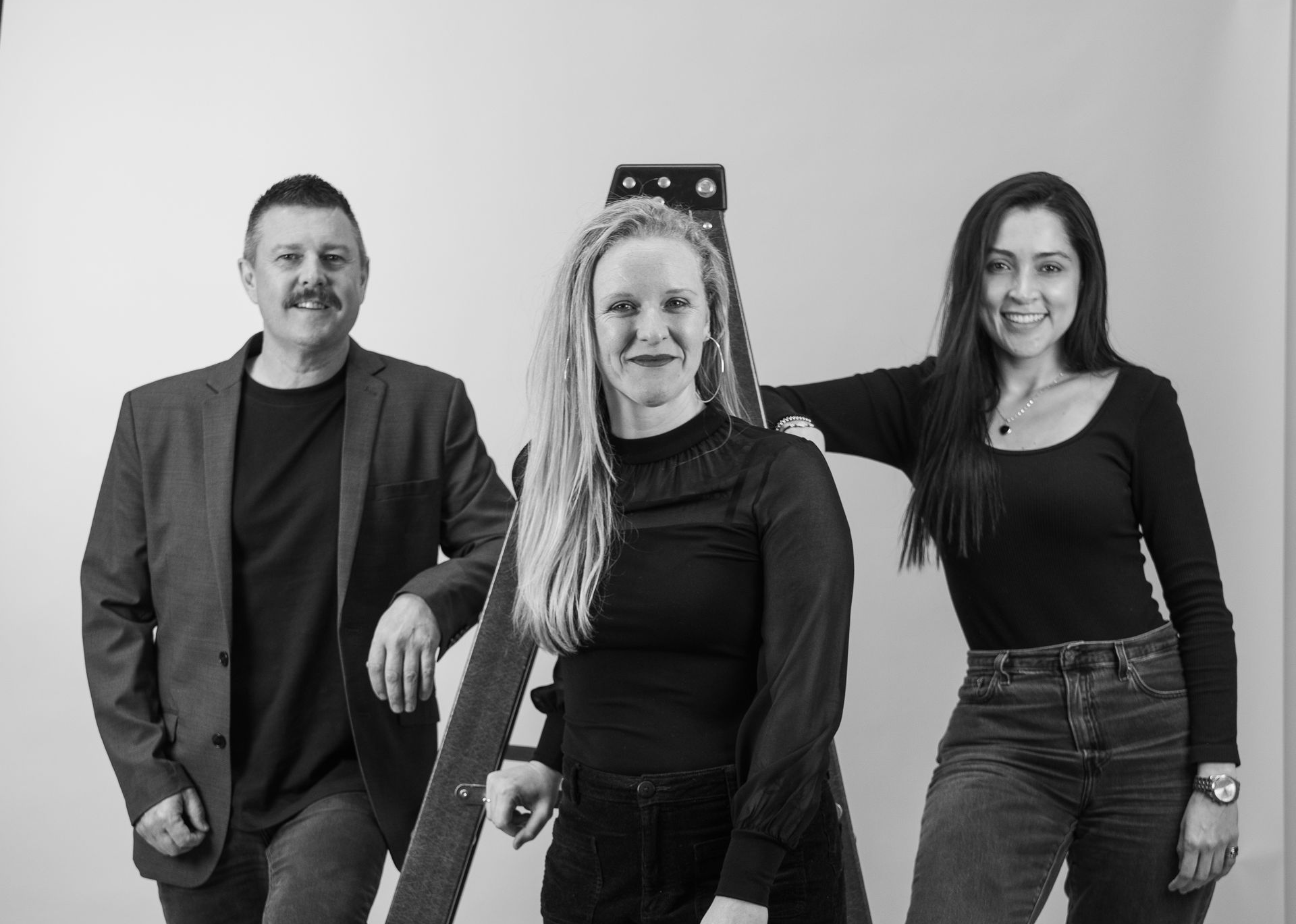Three people pose near a ladder. Woman in middle stands, others lean on ladder. Studio setting, black and white — Gracia Building Design In Kiama, NSW