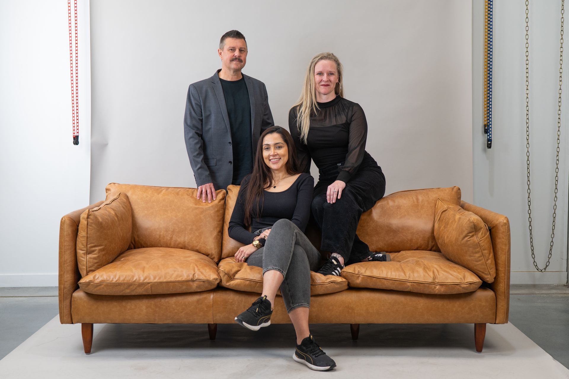 Three people pose on a brown leather couch against a white wall. A woman sits centred, two stand behind her — Gracia Building Design In Kiama, NSW