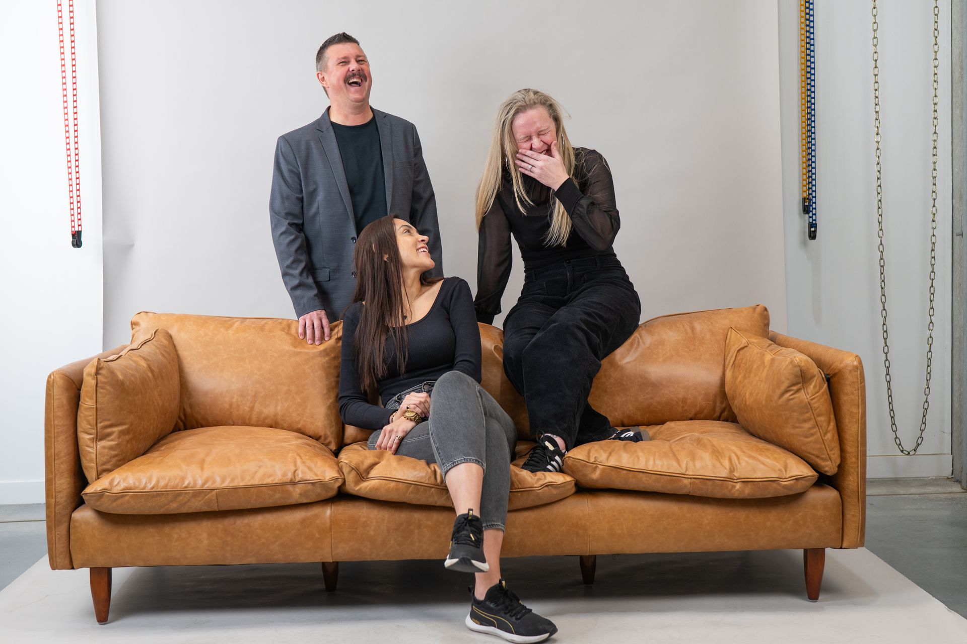 Three laughing people on a brown leather couch. One woman sits, another leans over laughing, and a man stands behind. White wall background — Gracia Building Design In Kiama, NSW