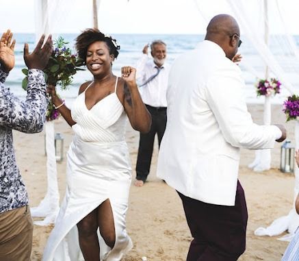 Bride and groom dancing at beach wedding ceremony. Guests celebrate, arch and flowers visible.