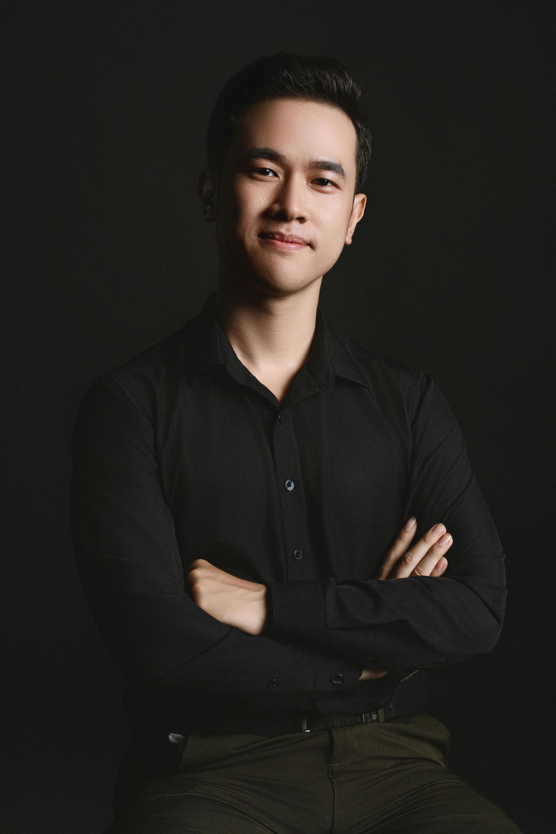 Man in black shirt with arms crossed, smiling. Dark backdrop.