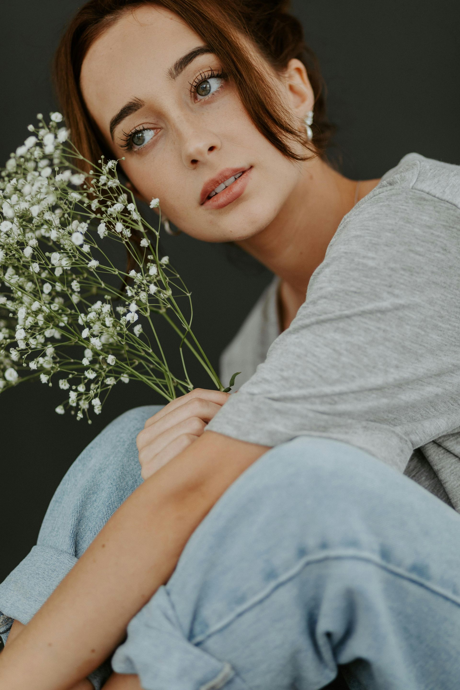 Woman with light brown hair, holding white flowers, looks pensively to the side. Gray shirt and blue jeans.