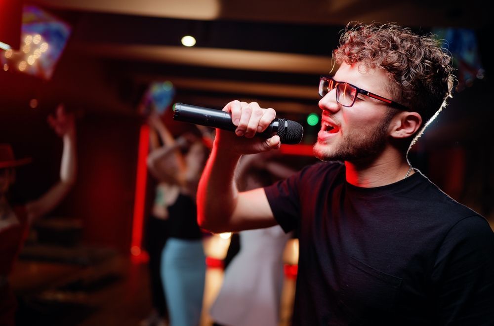 Man singing into a microphone at a karaoke bar, red lighting, people in the background.