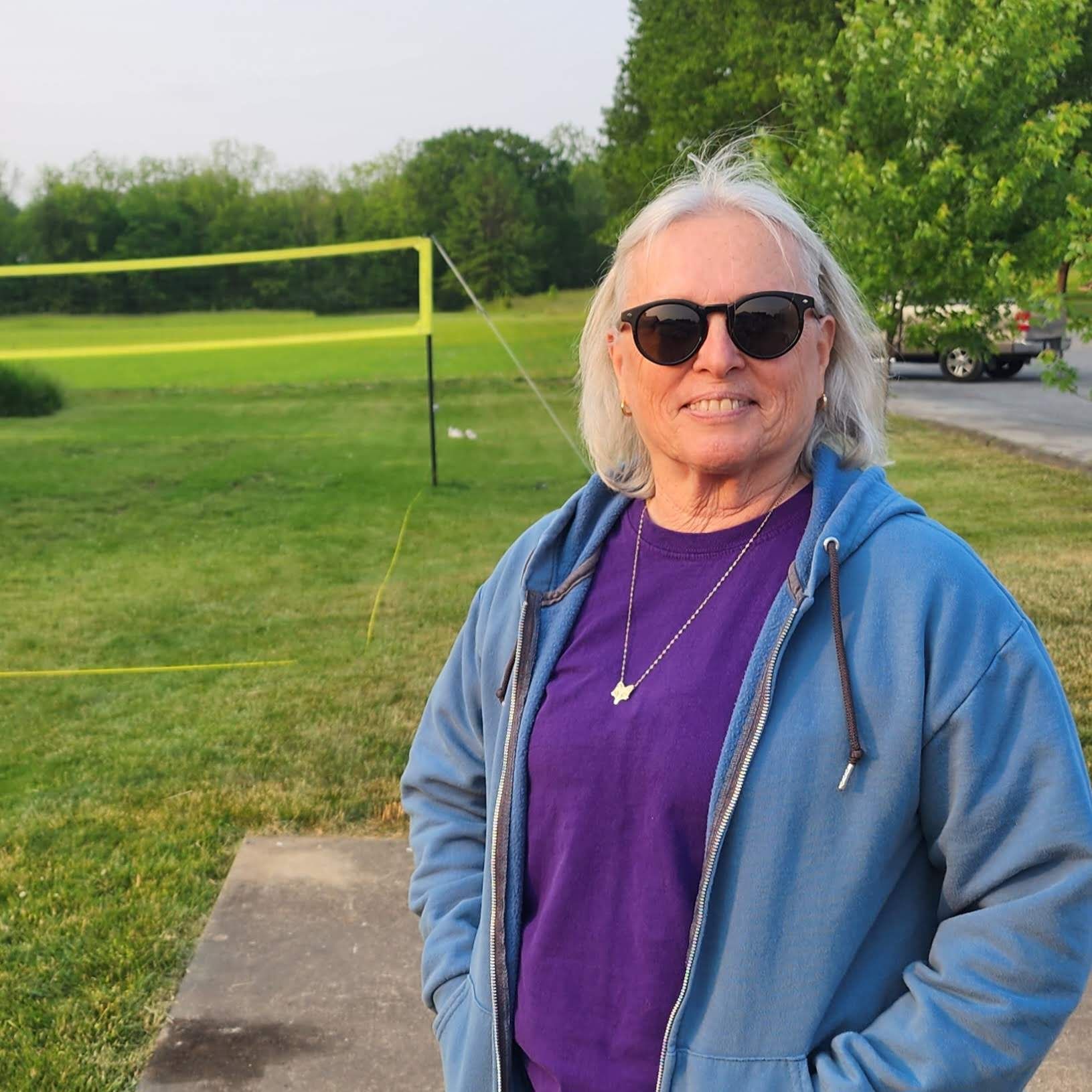 A woman wearing sunglasses and a blue jacket is standing in front of a volleyball net