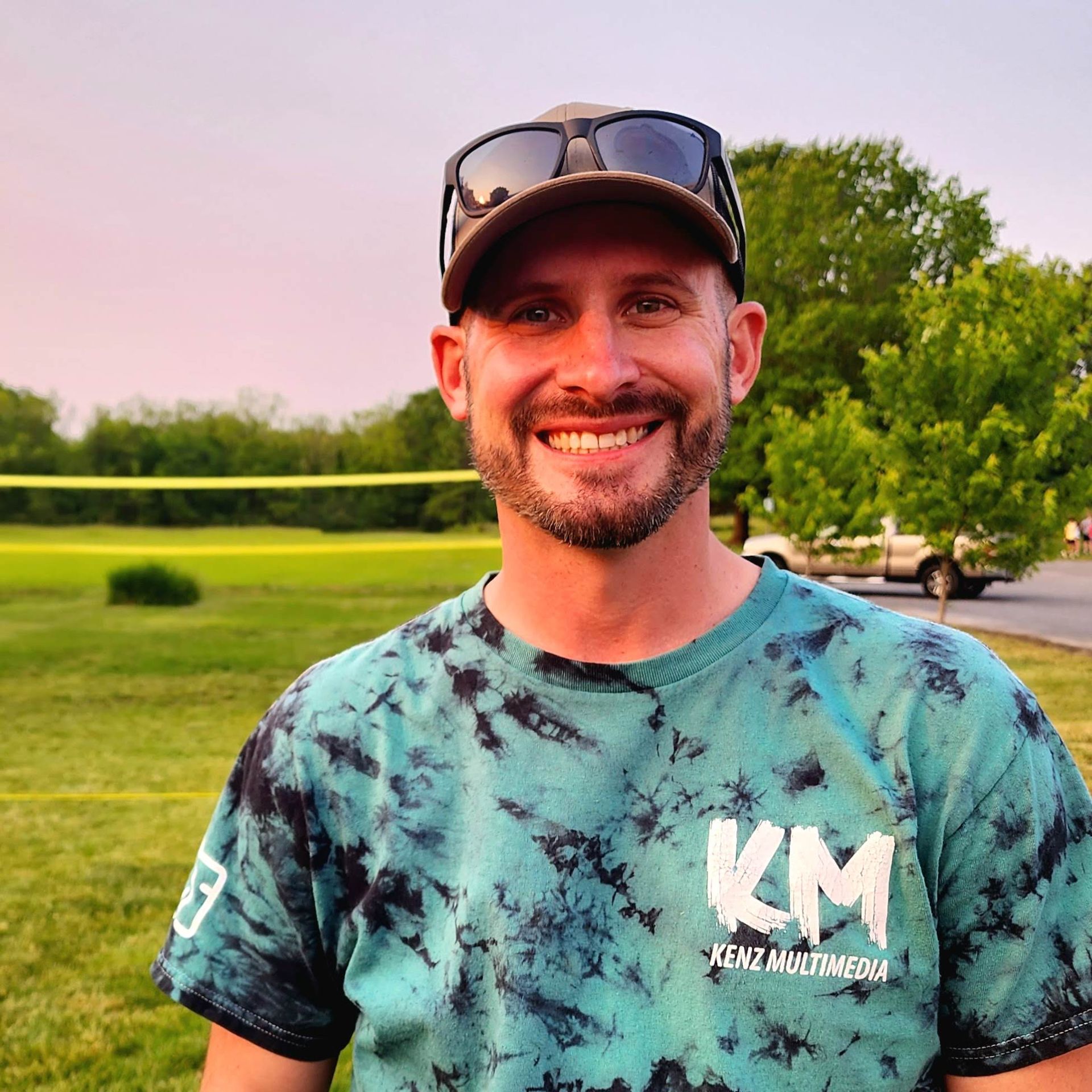 A man wearing sunglasses and a km shirt smiles for the camera