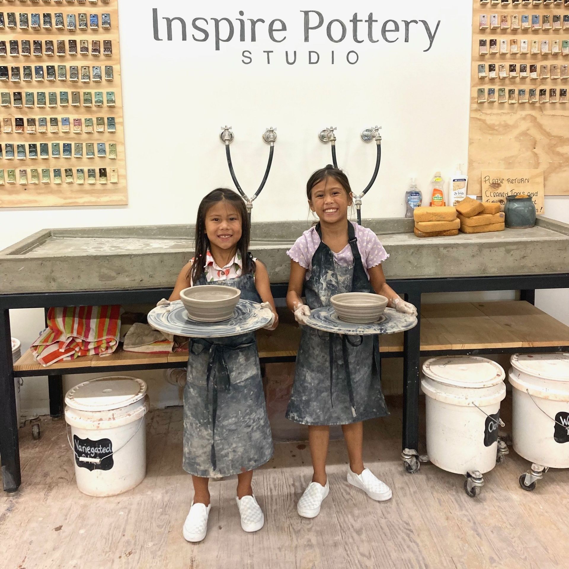 two young girls holding bowls in front of a wall that says inspire pottery studio