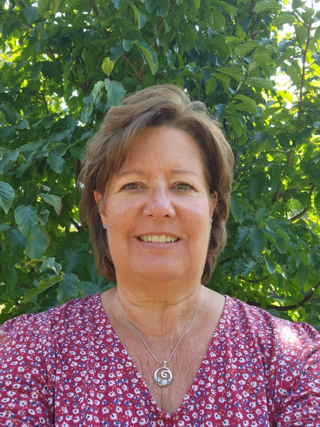 A woman in a red shirt and necklace is smiling in front of a tree.