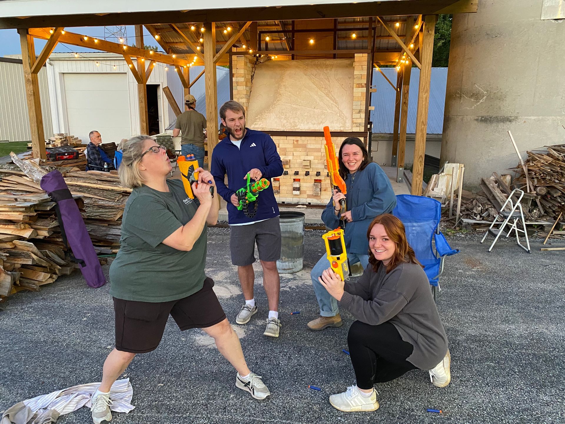 Four people posing with toy guns, outdoors near a stone structure. Two are kneeling, smiling, and aiming.