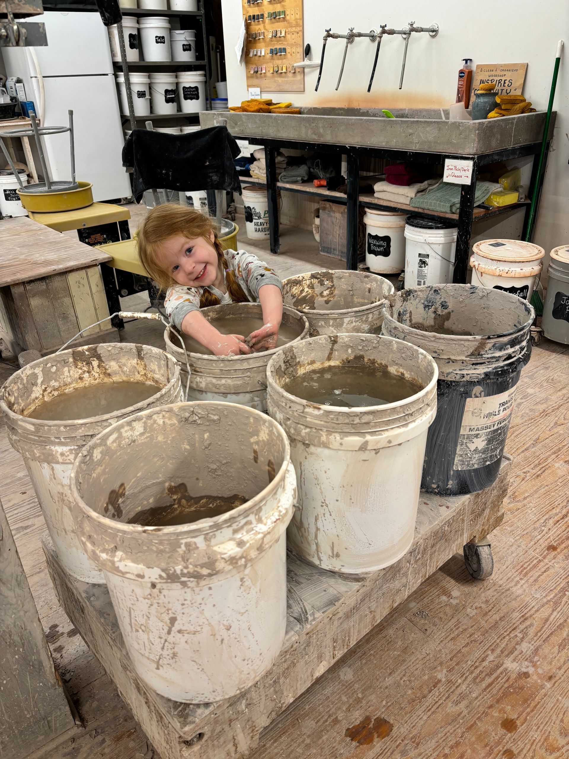 A little girl is playing in buckets of mud in a workshop.