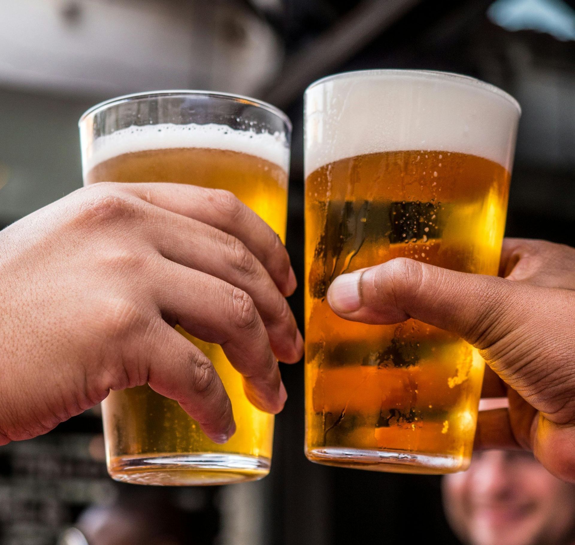 Two hands clink beer glasses together, filled with golden liquid, outdoors.