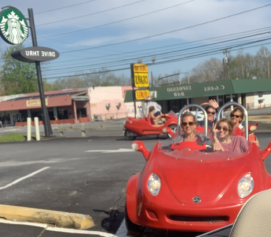 People in red novelty cars at a Starbucks drive-thru, waving with buildings in the background.