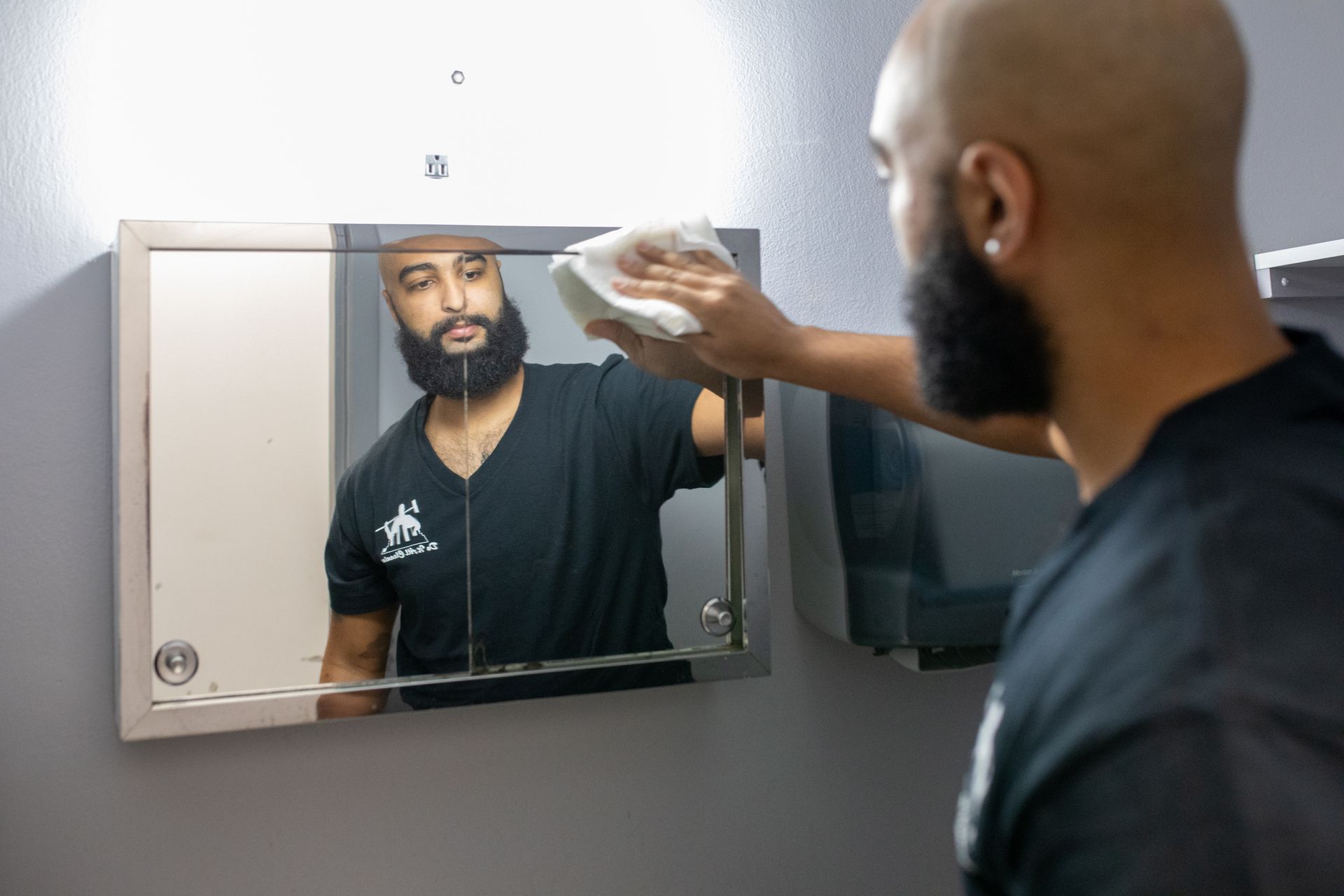 Man wiping a mirror in a bathroom with a white cloth; water droplets visible.