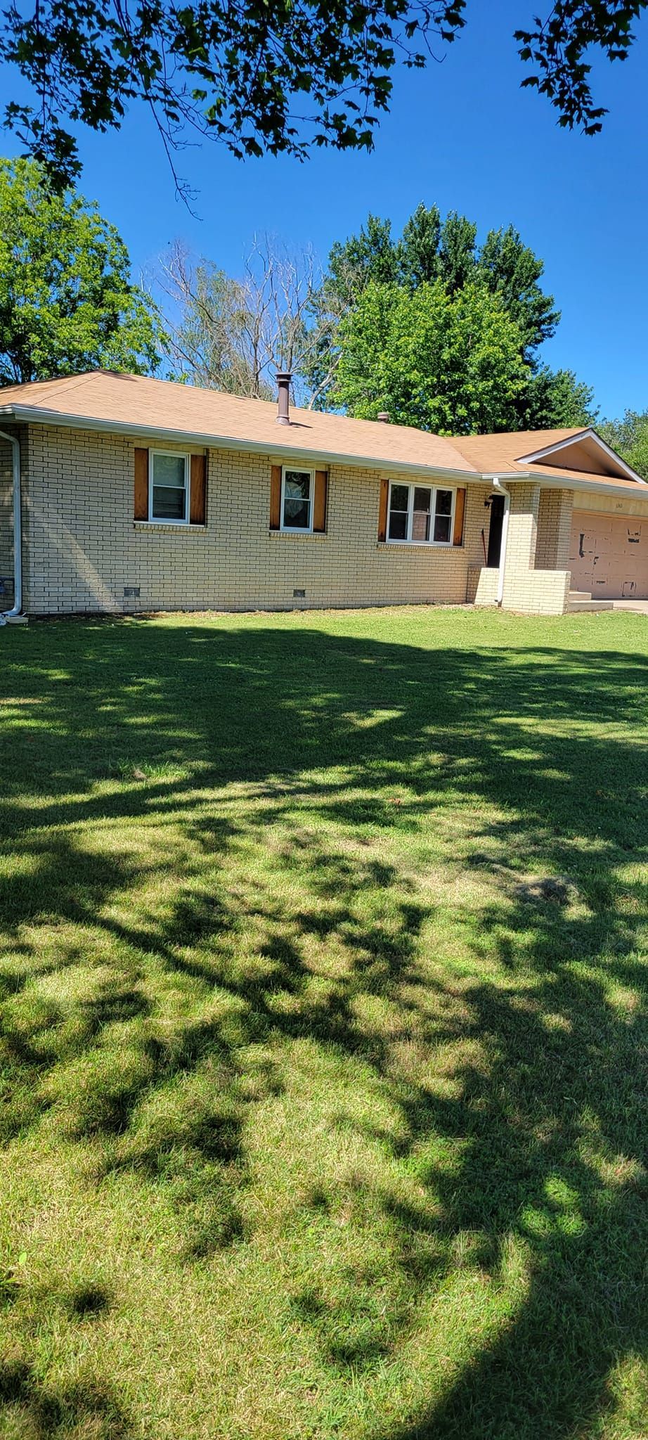 A beige ranch-style house with a tan roof, brown shutters, and a green lawn under a bright blue sky.