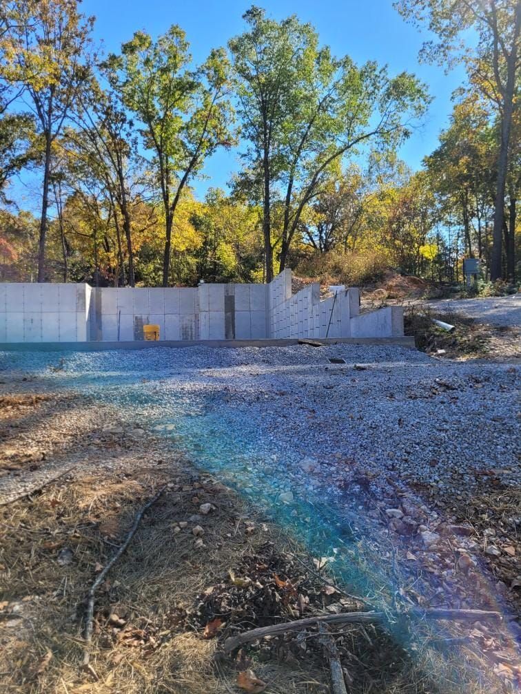 Construction site with concrete foundation walls in front of trees with fall foliage; gravel ground in foreground.