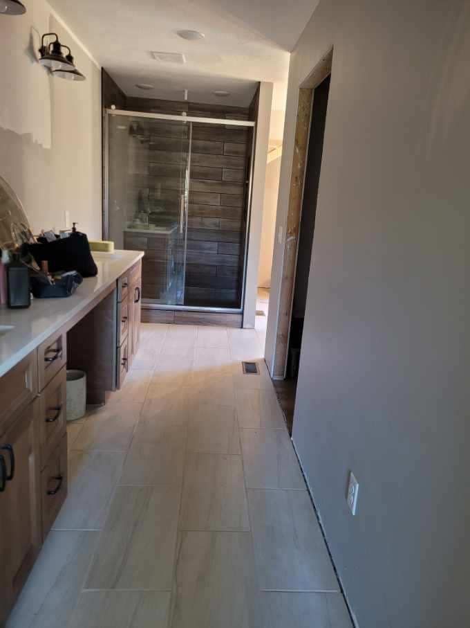 Bathroom with wood-toned cabinets, tiled floor, glass shower, and a doorway on the right.