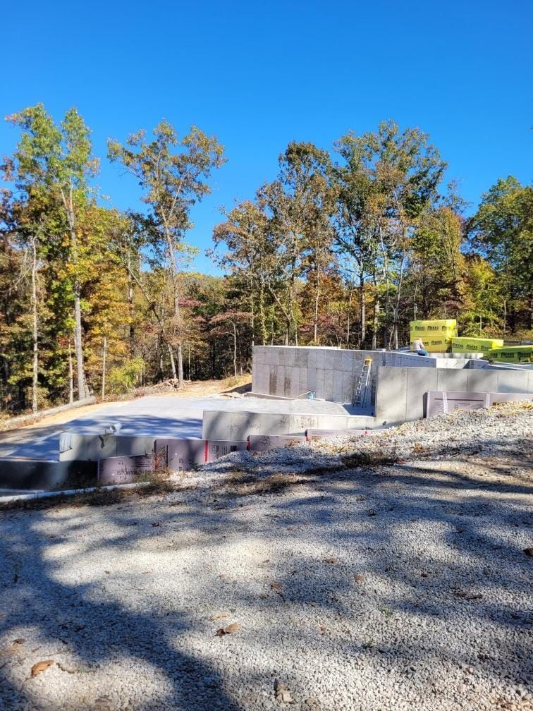 Construction site: concrete foundation and blocks, trees in background under blue sky.