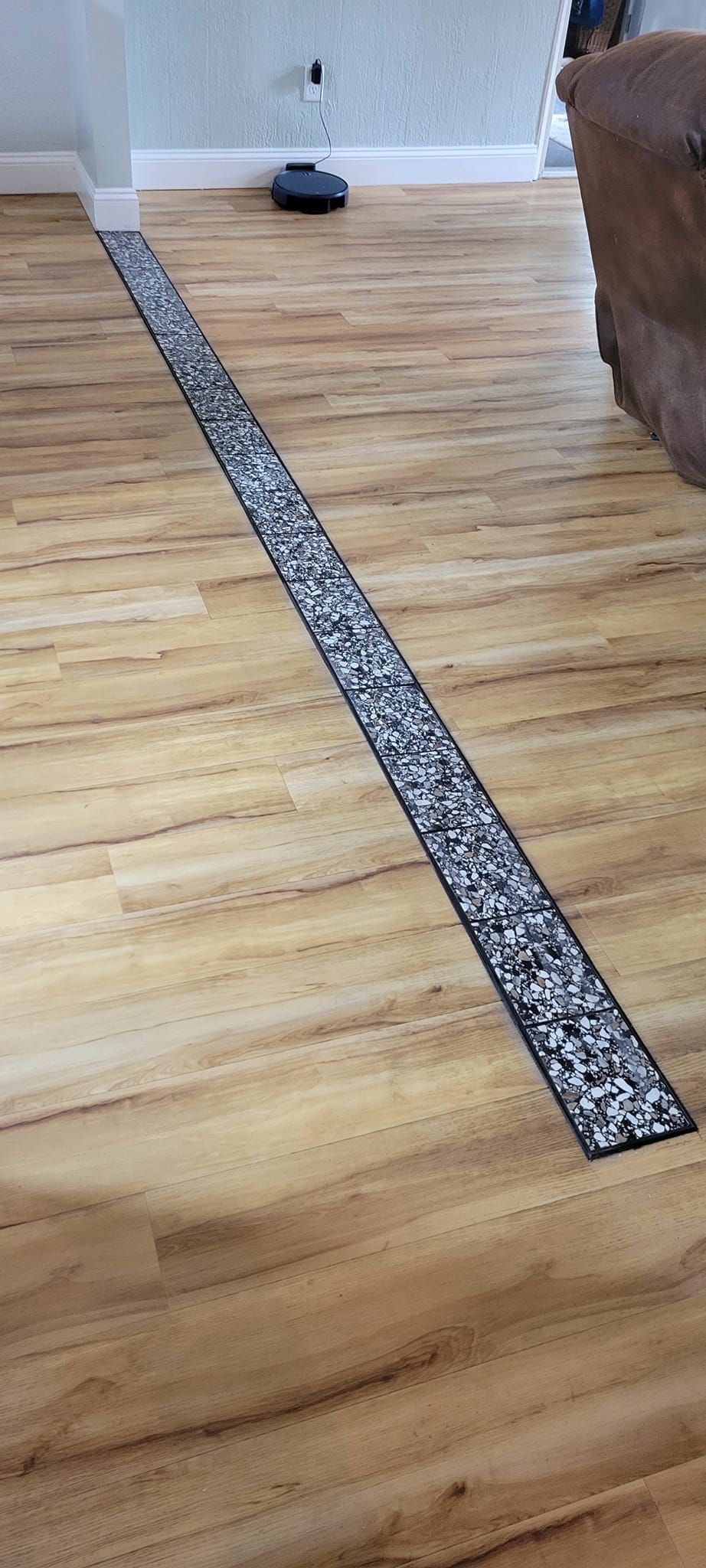 Wooden floor with a long strip of gray and black mosaic tile inset. A Roomba sits in the background.
