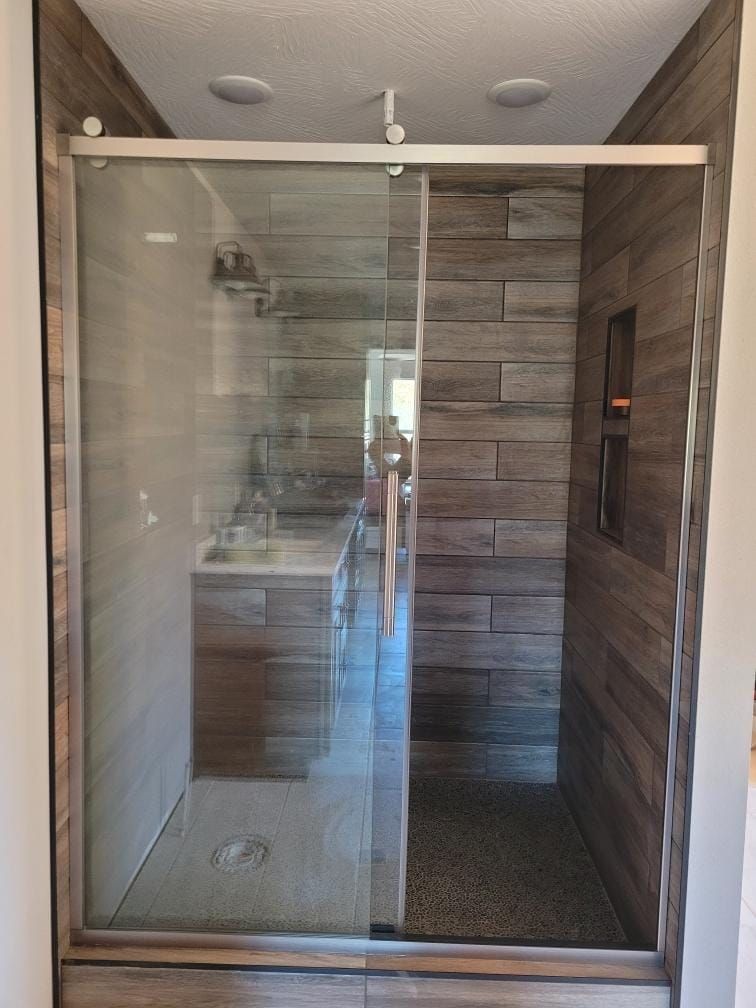 Shower with a sliding glass door, brown tiled walls, and pebble flooring.